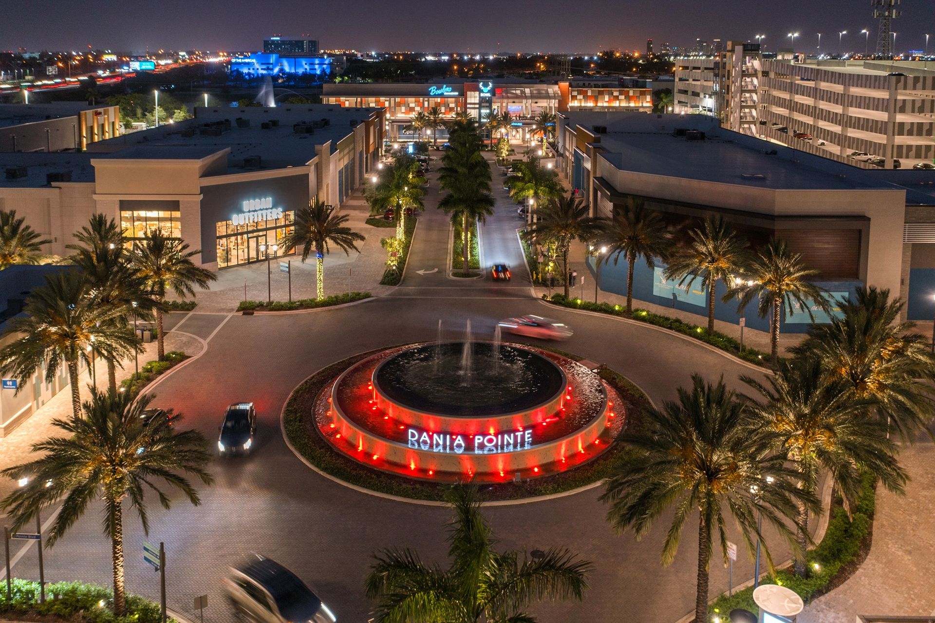 Dania Pointe fountain plaza at night, with the illuminated Dania Pointe sign, palm trees and lit storefronts